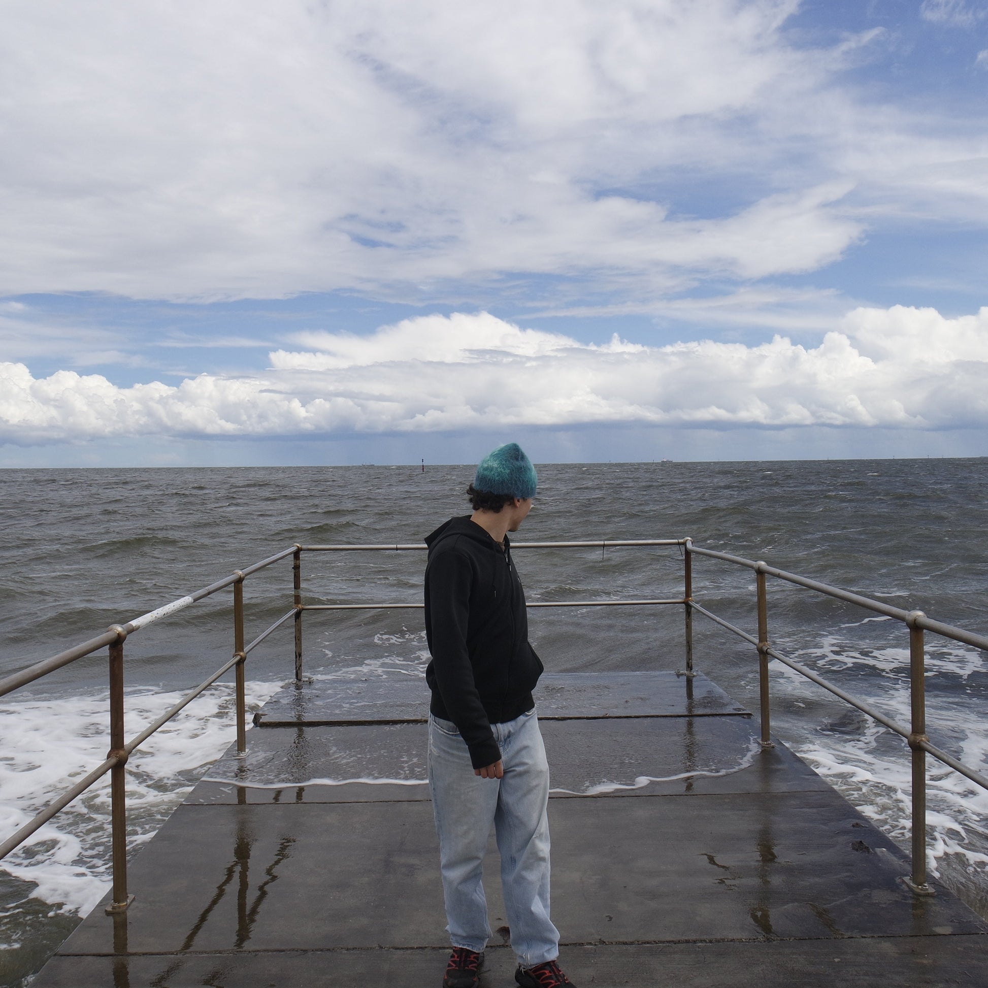 Person standing on a pier looking out at the ocean with a cloudy sky.